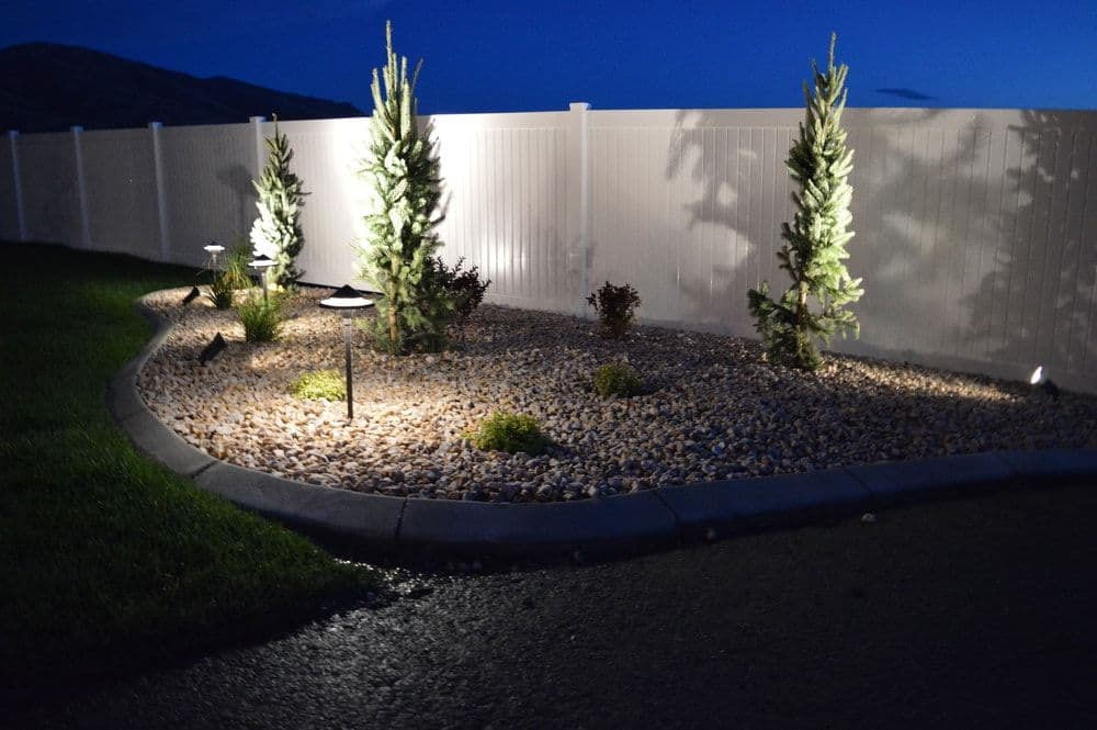 Illuminated landscape with stones and shrubs beside a white fence at night.