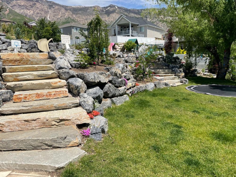 Stone steps lead through a landscaped garden with flowers and a view of mountains and homes.