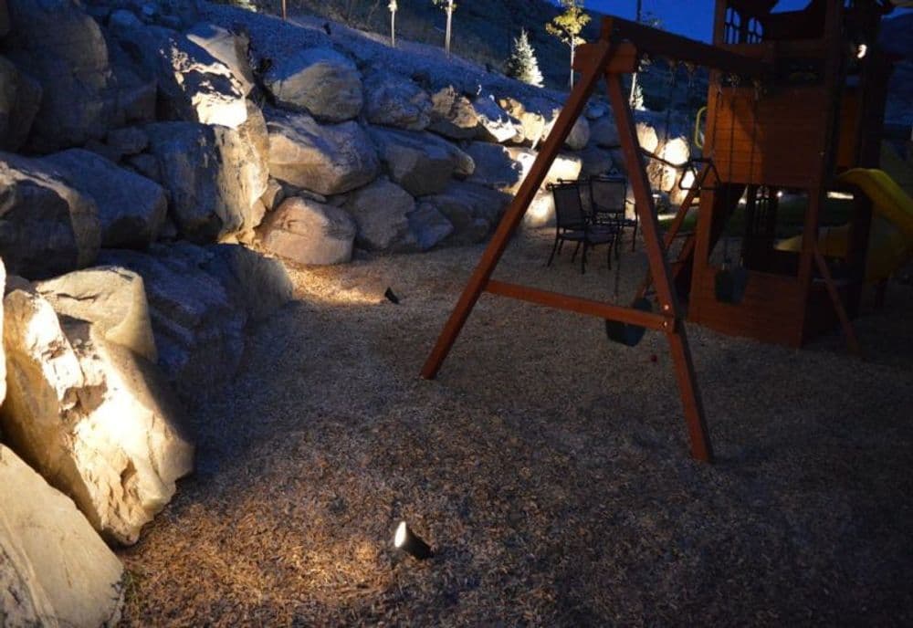 Playground with swings and slide illuminated by lights at night, surrounded by rocky terrain.